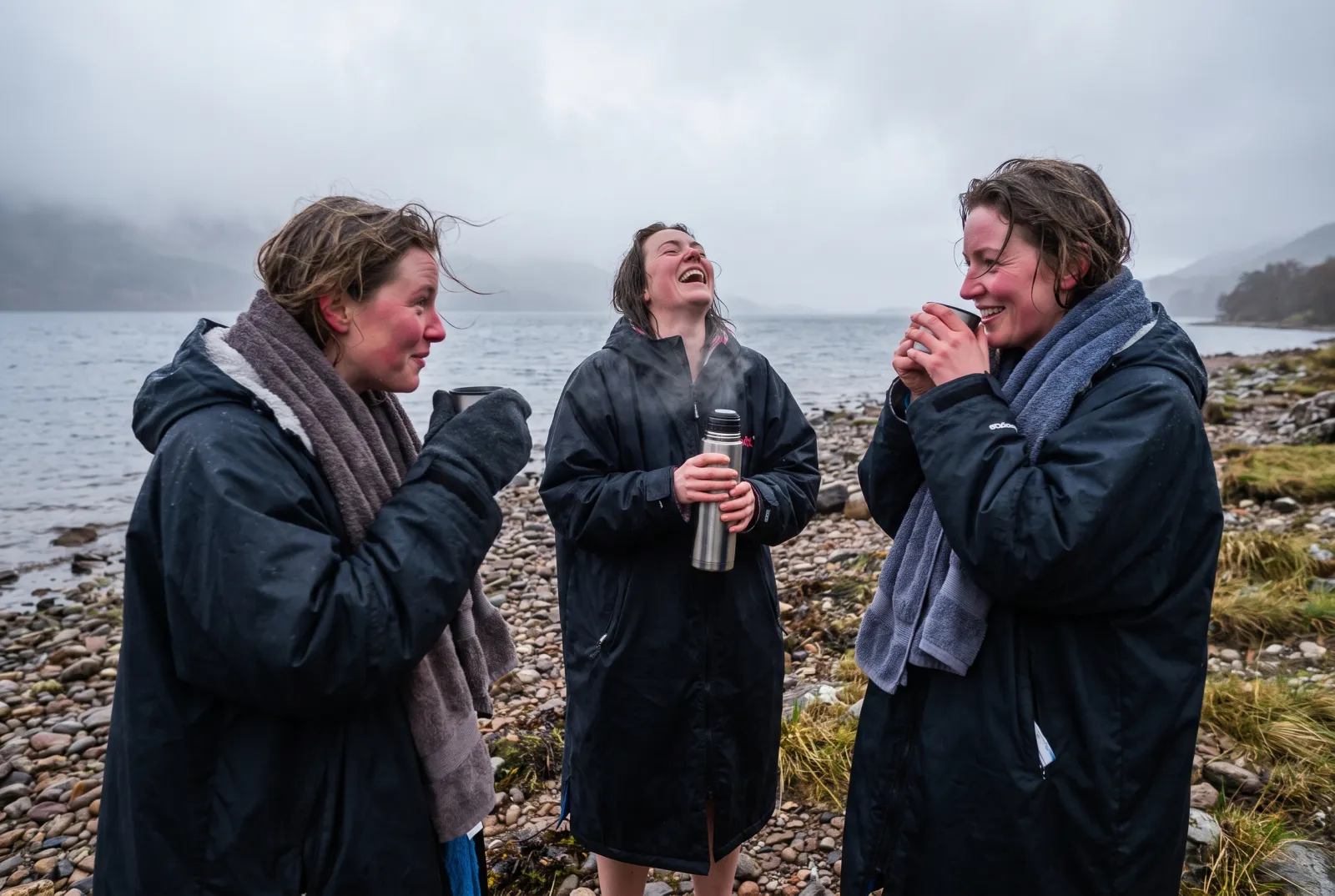 Three women in long swim robes laughing on a pebble shore after a cold loch swim, mist on the hills behind