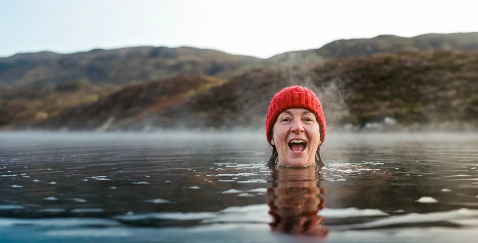A smiling woman in a red woolly hat up to her shoulders in a misty loch, heather-brown hills behind her